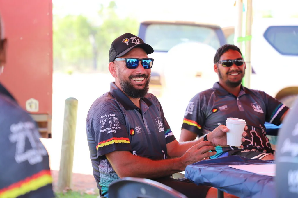 Two men wearing KAMS "Get your 715 health check today" shirts smiling and talking during an outdoor community event in the Kimberley.