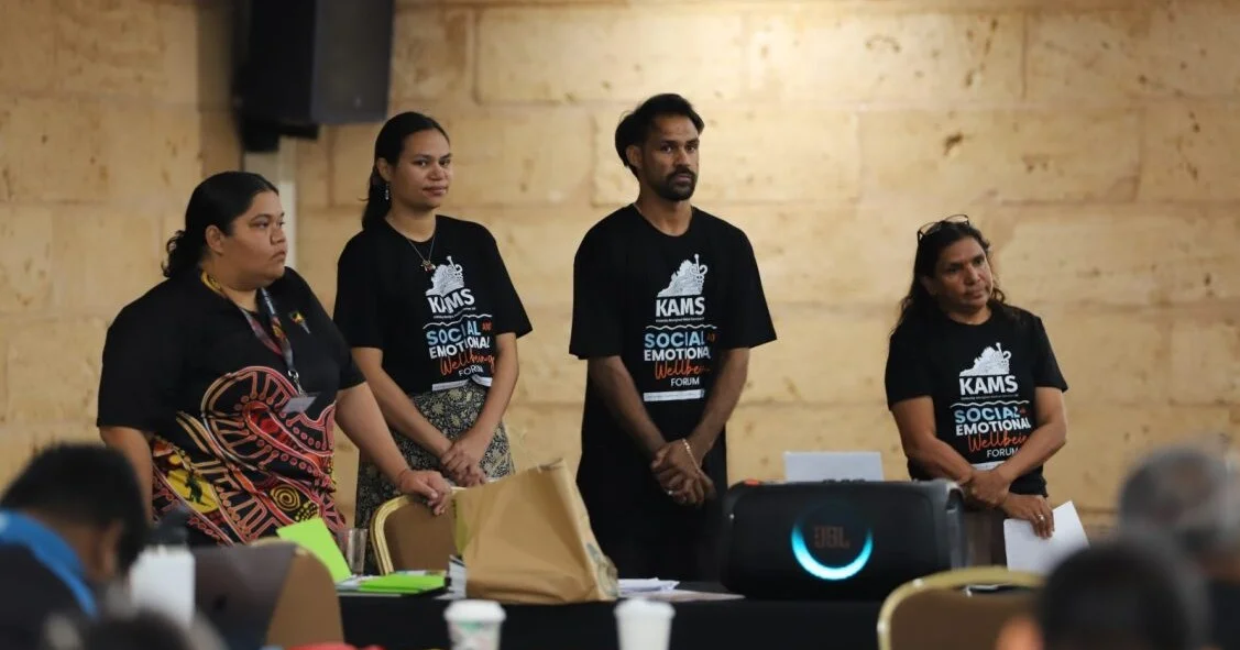 Four Aboriginal health staff members wearing matching KAMS Social and Emotional Wellbeing Forum t-shirts standing around a table at a community workshop.